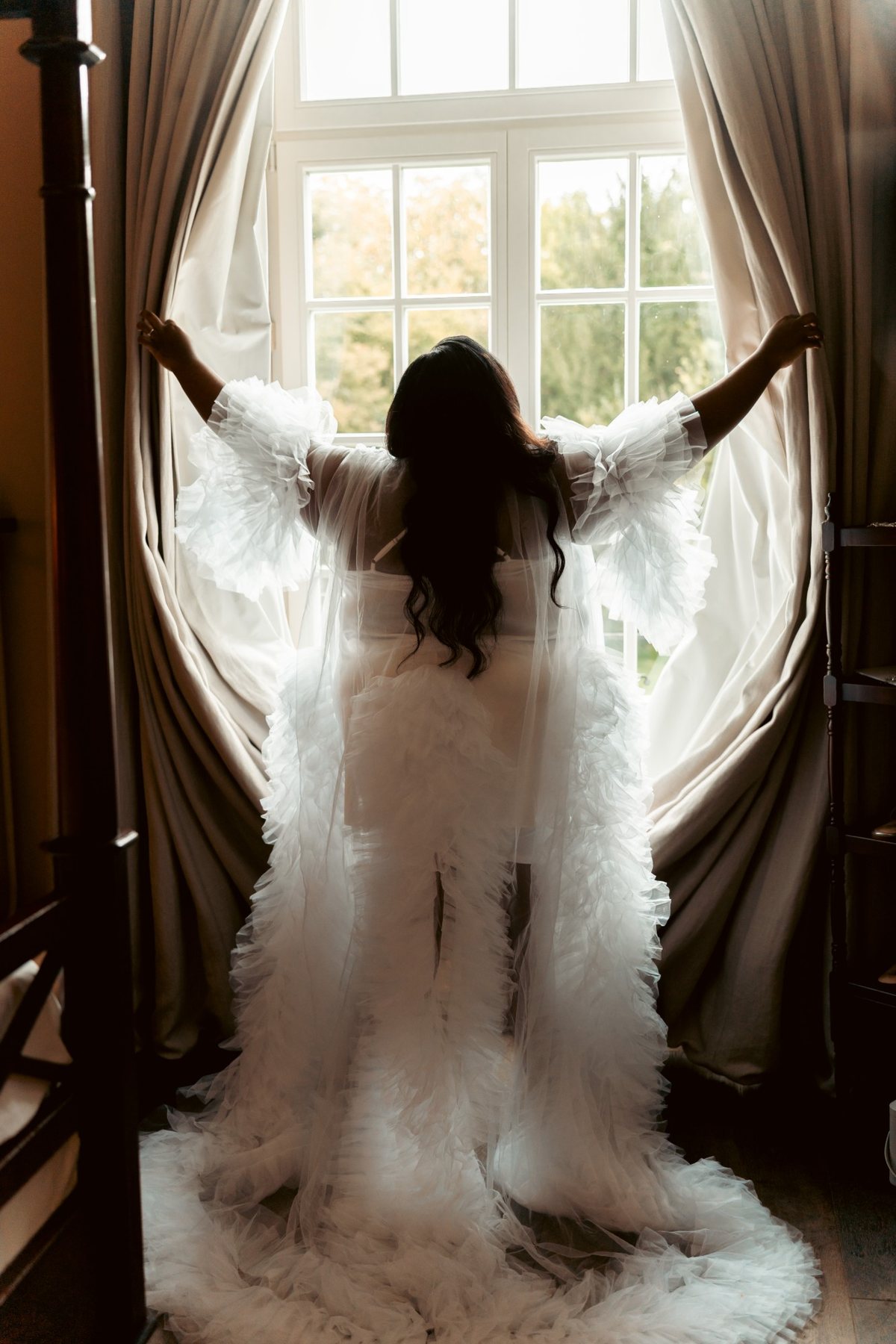 Une mariée en robe de tulle ouvre les rideaux de sa chambre